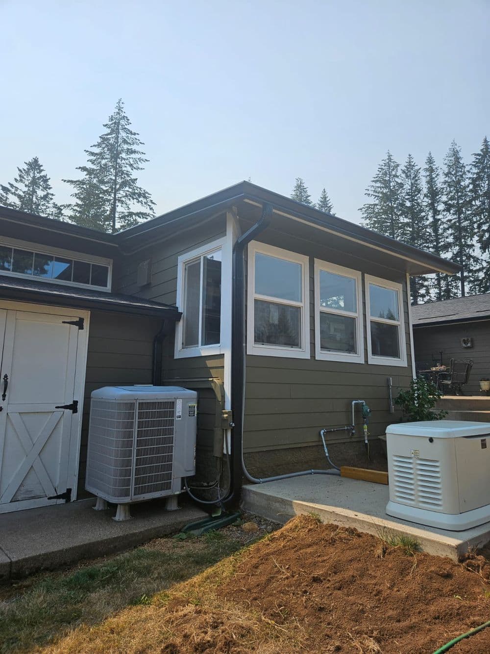 Modern home exterior with large windows, outdoor unit, and landscaped yard in a forested area.