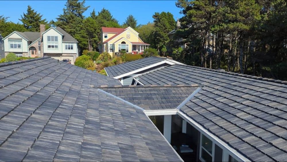Aerial view of modern roofs with gray shingles, surrounded by lush greenery and homes.