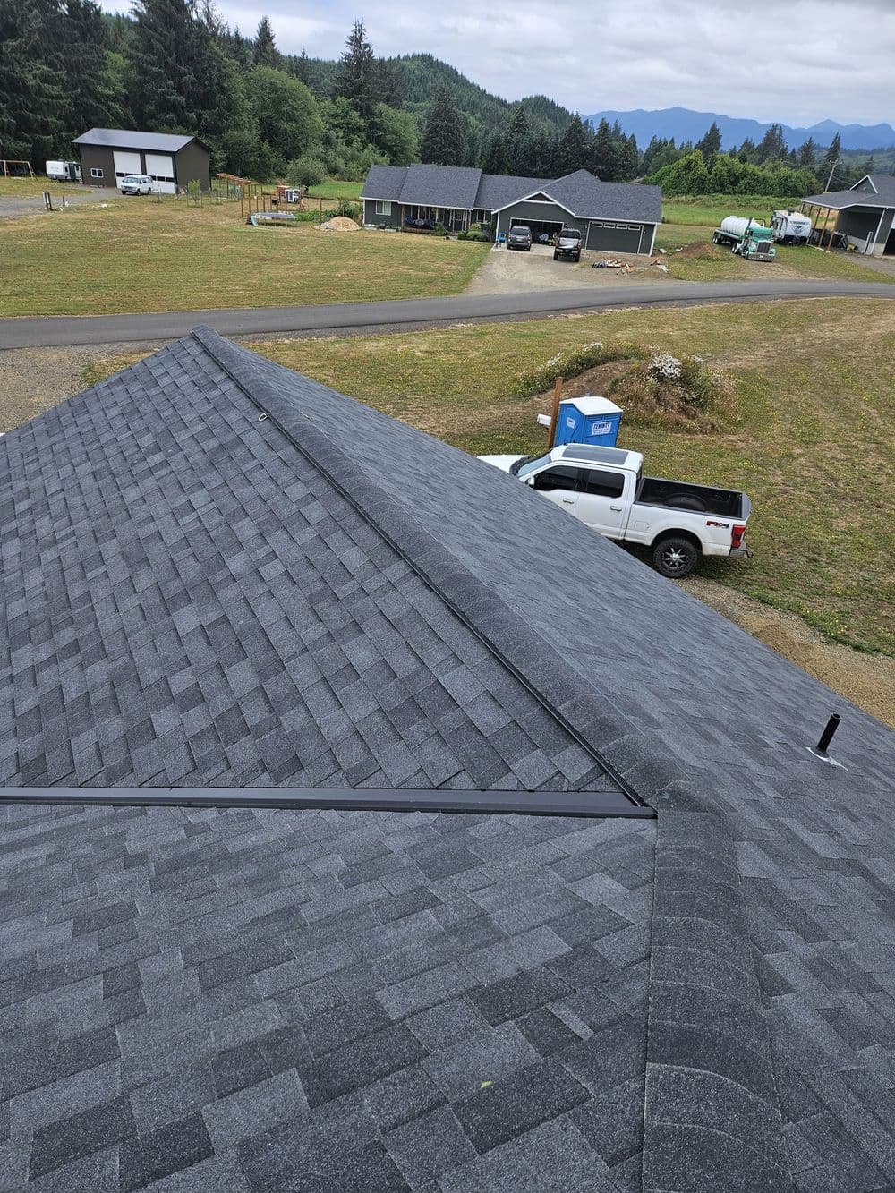 Aerial view of a gray shingle roof on a house with green landscape and distant mountains.