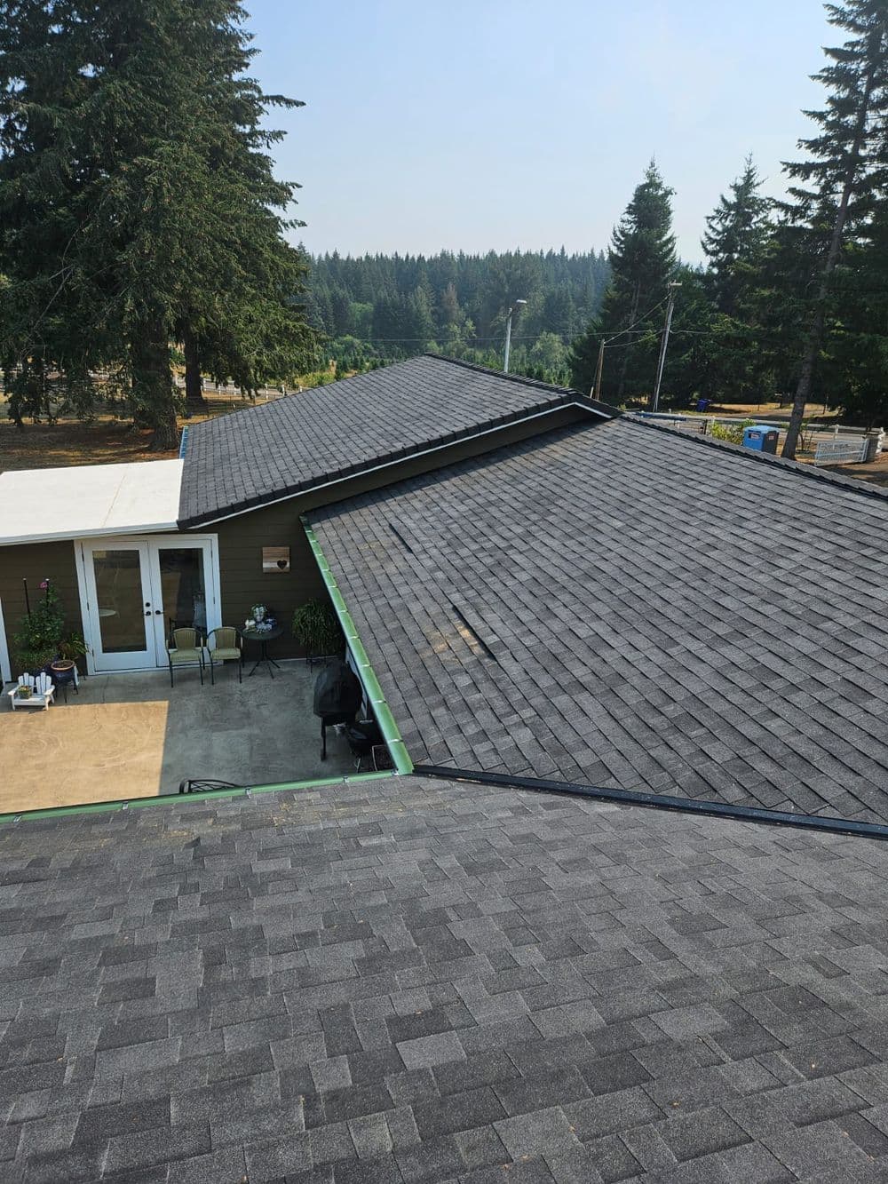 Aerial view of a gray shingle roof on a house surrounded by tall trees and open space.