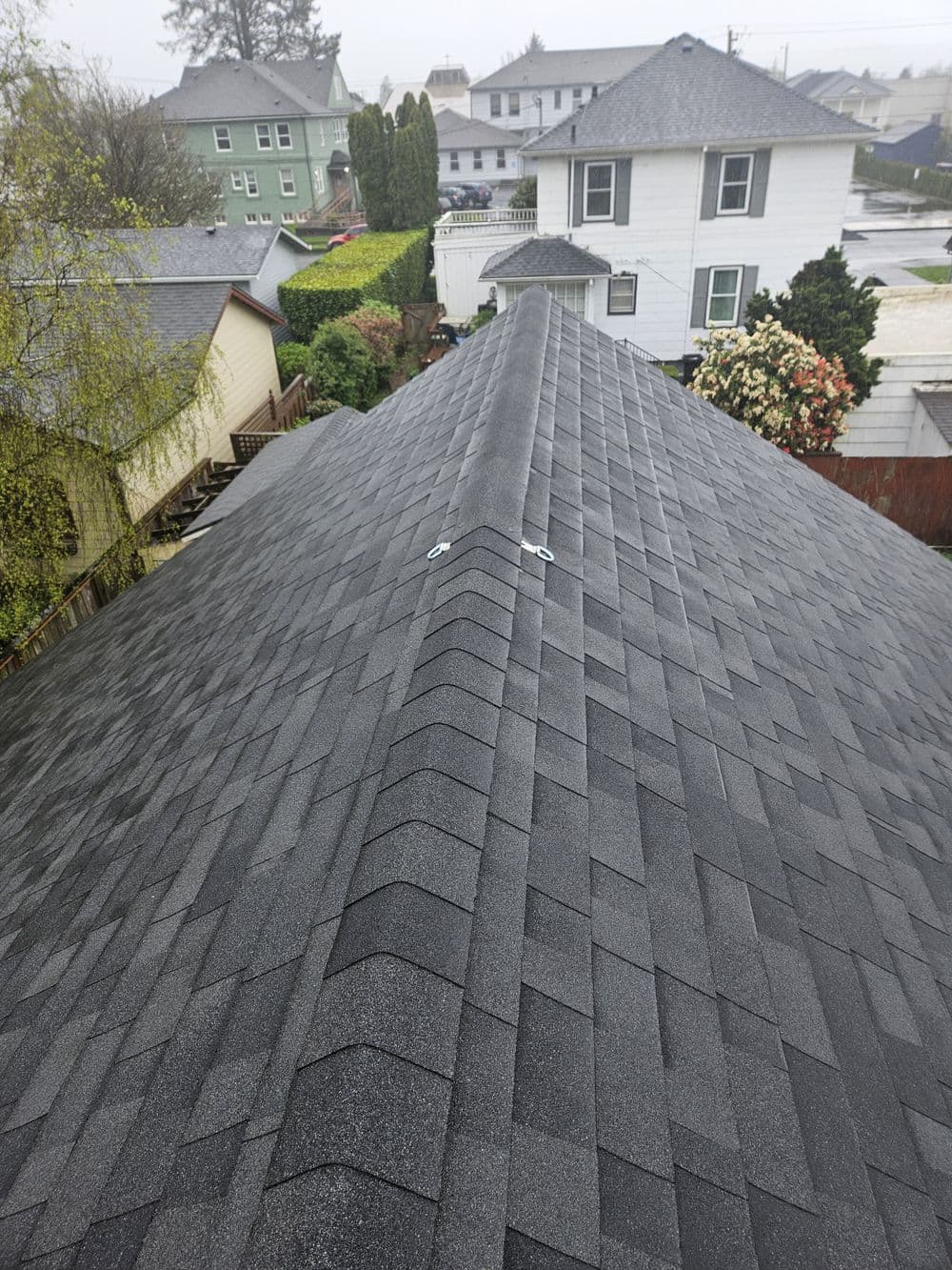 Aerial view of a grey shingle roof with residential homes and greenery in the background.