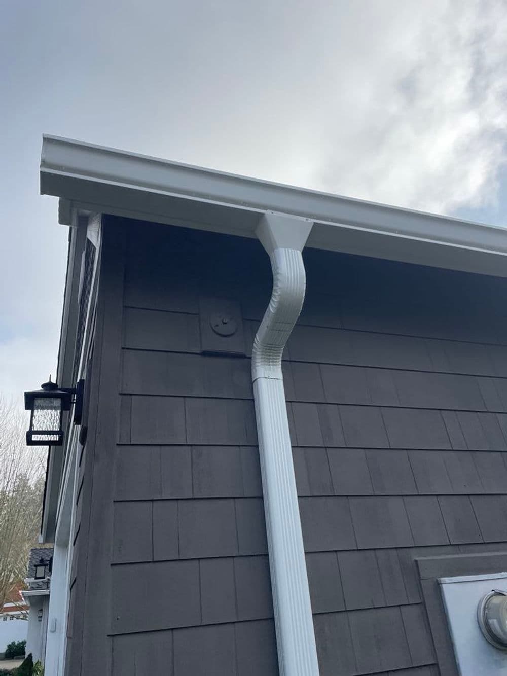 Gutter system and downspout on the side of a house with gray siding and cloudy sky.