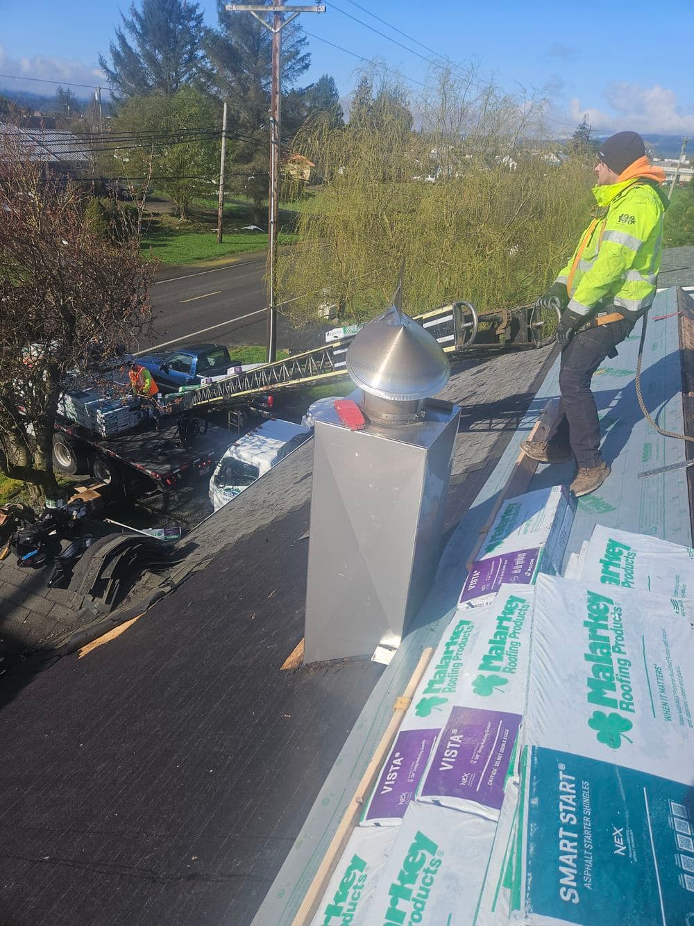 Roofer inspecting chimney cap installation on residential roof with truck and ladder below.