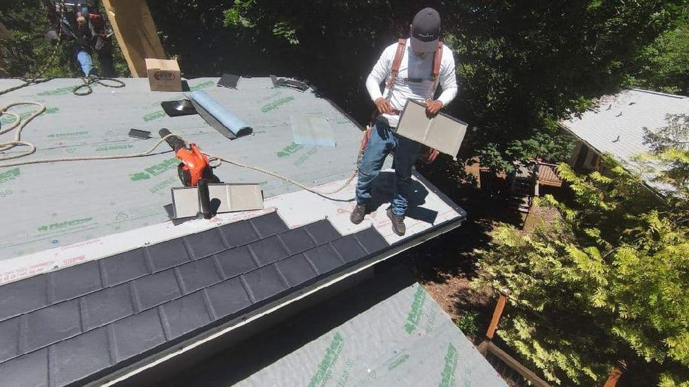 Worker installing roof shingles on a residential home with tools and materials visible.