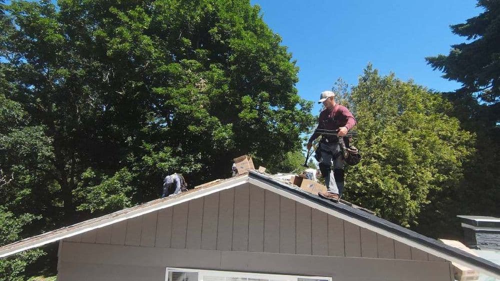 Roofer installing shingles on a house under a clear blue sky surrounded by trees.