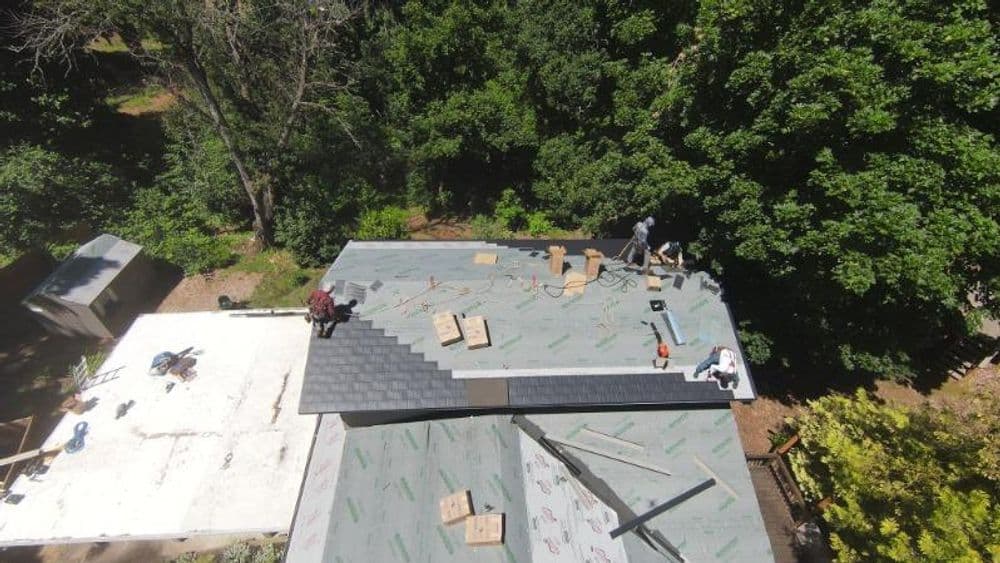 Roofing project in progress with workers installing shingles in a wooded area.