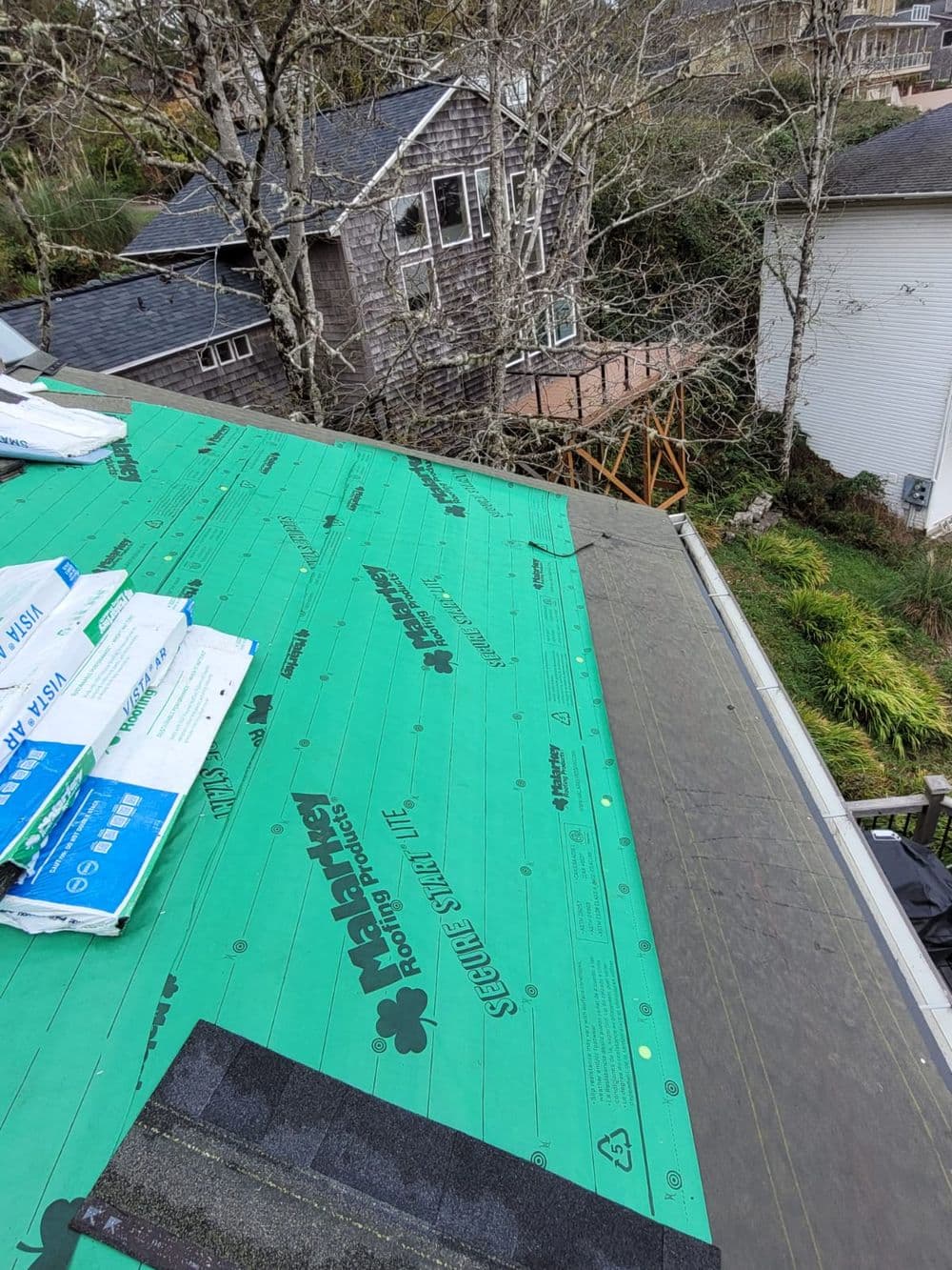 Roofing installation with green underlayment on a house, surrounded by trees and structures.