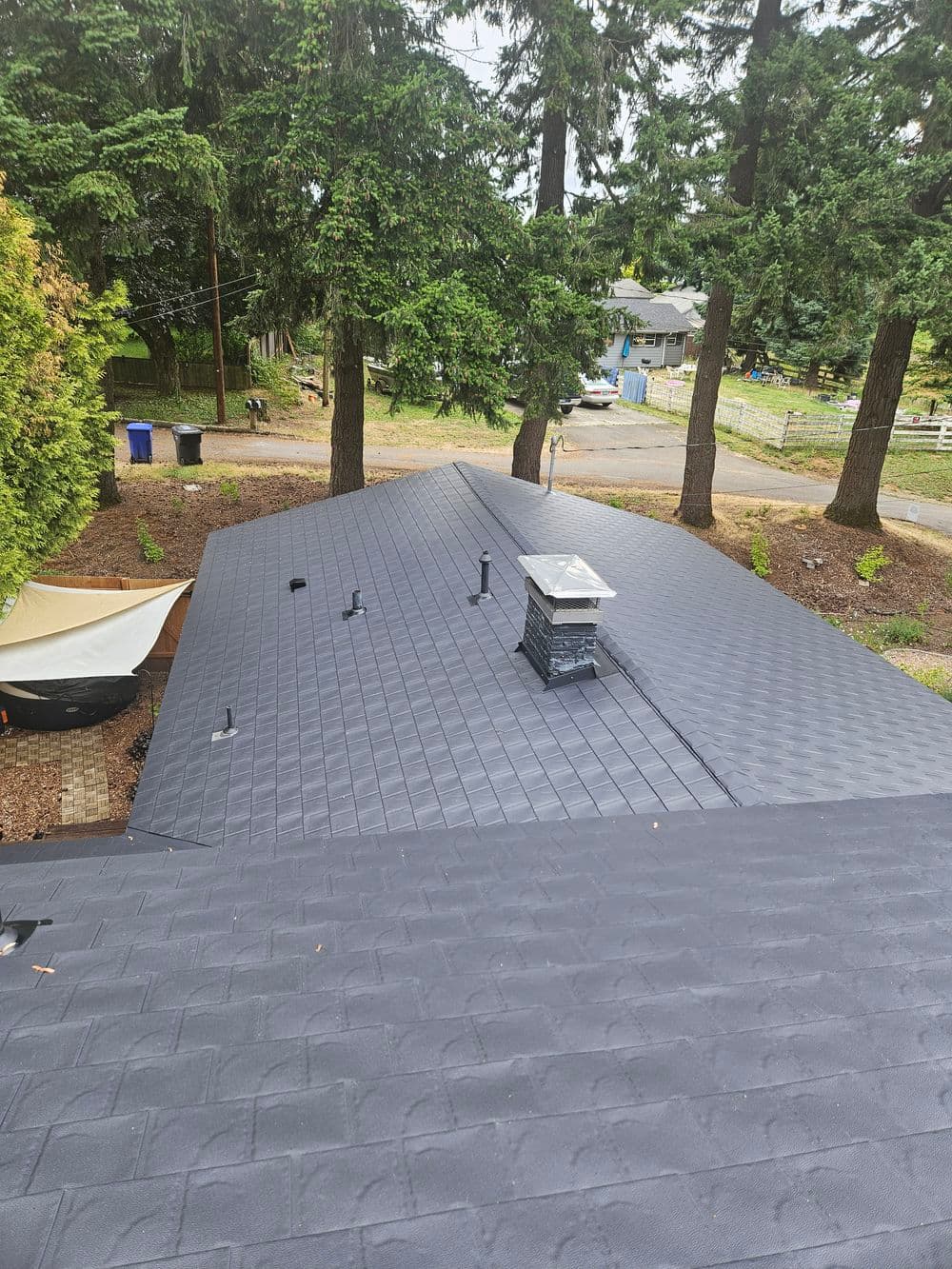 Aerial view of a newly installed dark gray roof with chimney surrounded by trees.